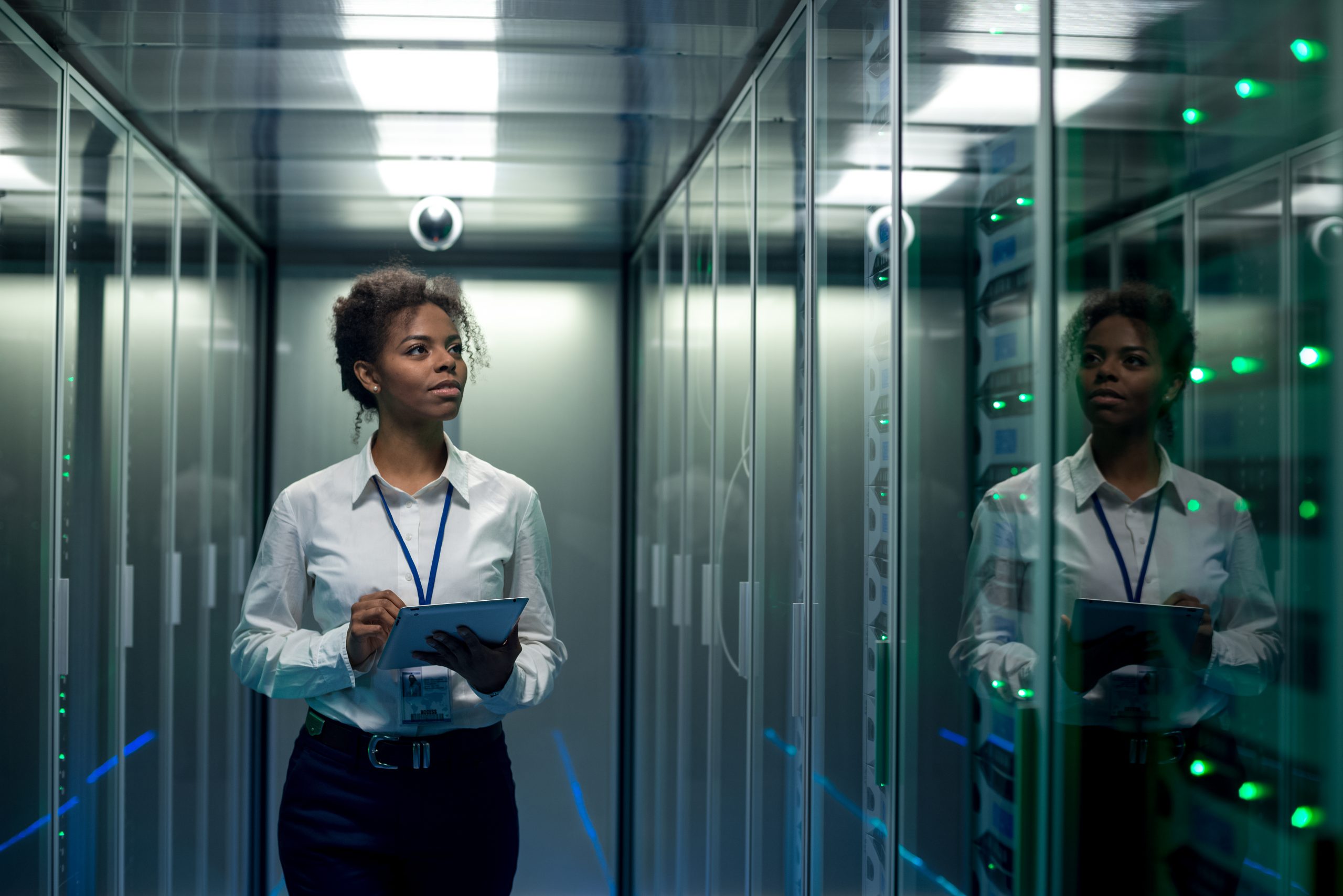 Femme afro-américaine utilisant une tablette en marchant dans le couloir du centre de données et en vérifiant le matériel sur la carte mère des racks de serveur